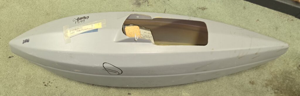 Gray fiberglass boat hull shell with an open cockpit cutout and taped labels on a carpeted floor.
