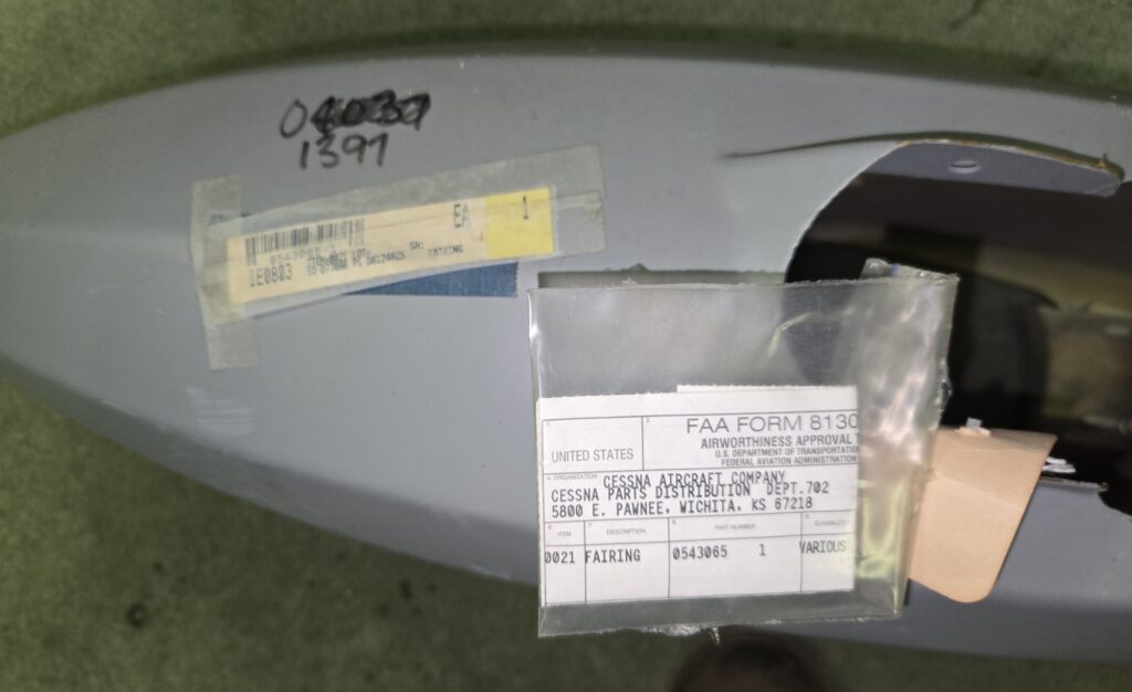 Close-up of a light gray aircraft surface with taped label and an FAA Form 8130 attached, containing serial numbers and Cessna part information.